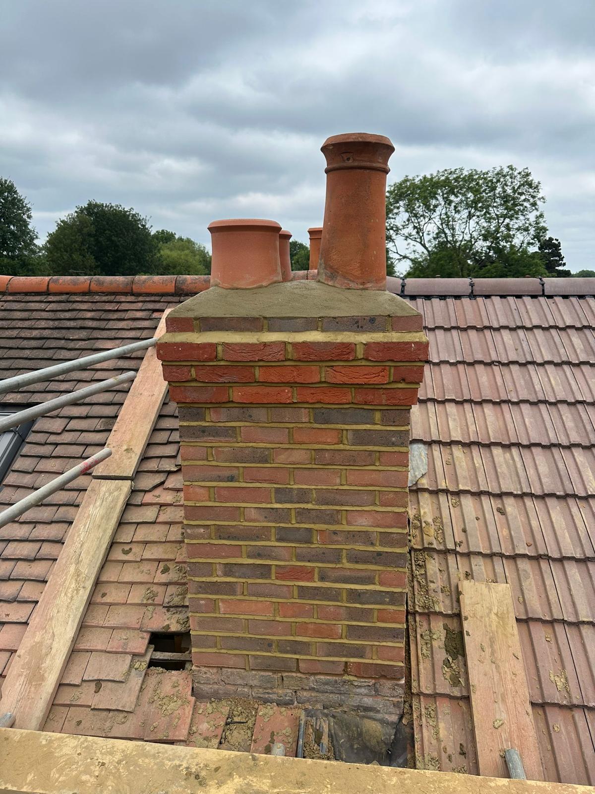 Repointed brick chimney with new terracotta pots on a clay-tile roof under repair